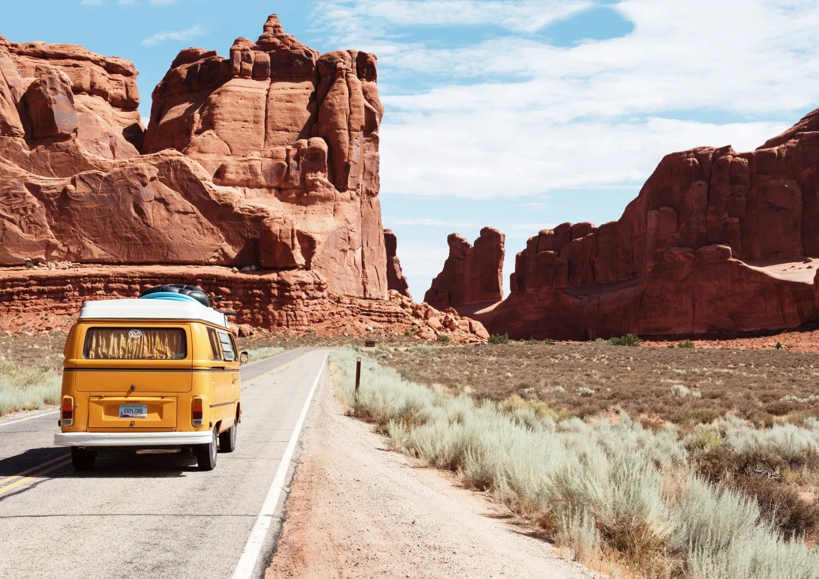 Yellow Volkswagen van on an empty road facing red rock landscapes, with an « EXPLORE » plate